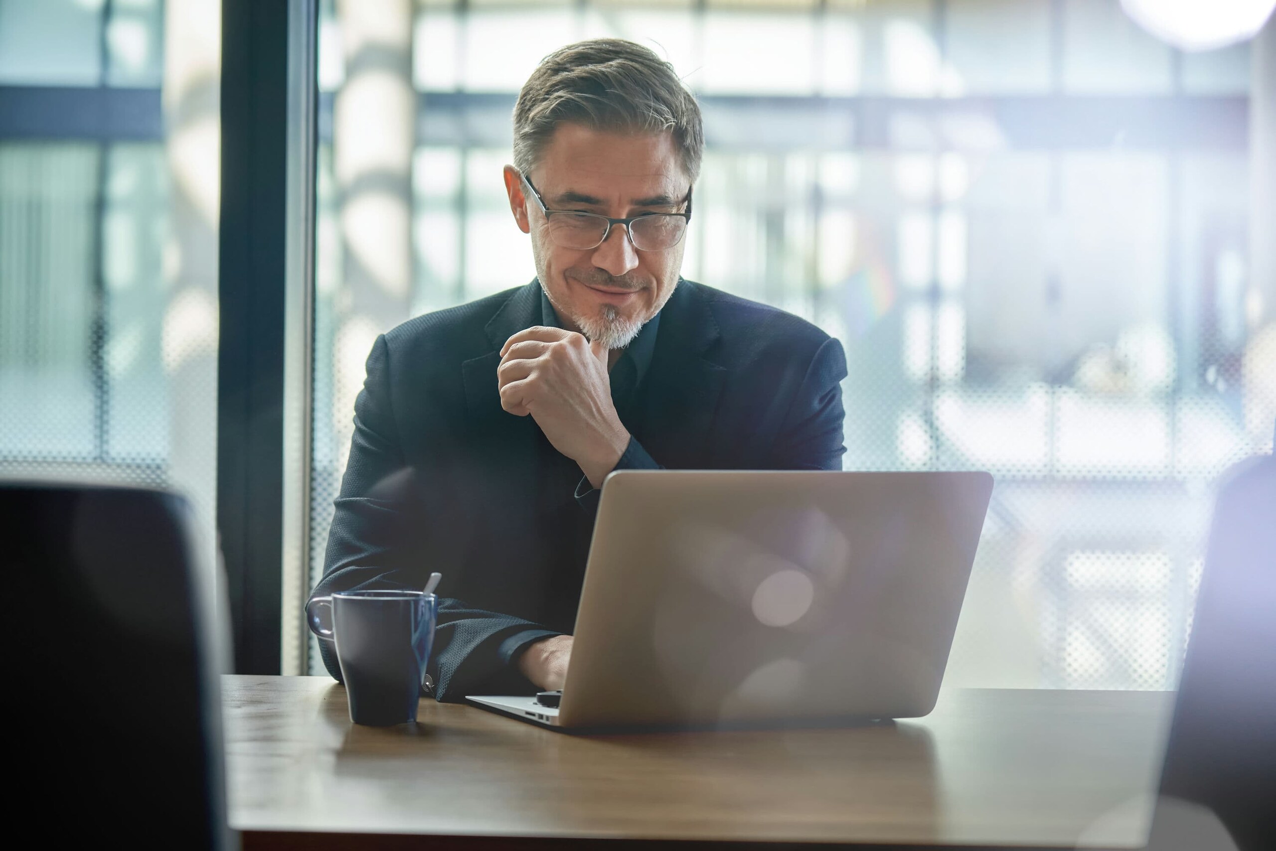A man wearing spectacles and working on a laptop, with a cup of coffee in hand